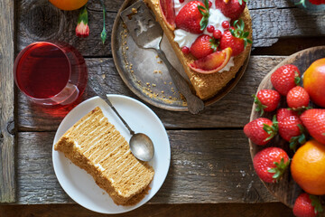 A piece of honey puff cake garnished with strawberries, red orange and cream. Flowers, wooden background, top view.