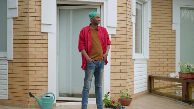 Smiling young African American man standing on porch waving to neighbors. Wide shot of positive happy relaxed guy resting at backyard on weekend. Lifestyle and leisure