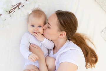 portrait of a mother with a baby, a mother kisses a child and gently hugs him on a white bed at home, maternal love and care