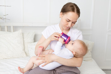 Obraz na płótnie Canvas mom drinks or bottle feeds a baby on a white bed at home, baby food concept
