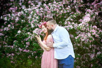 A young woman in a pink lace dress and a bearded adult man in a white shirt hold hands against the background of apple trees in bloom.
