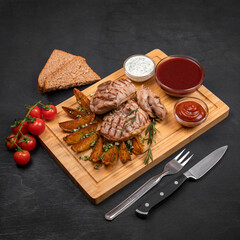 Grilled bbq meat steak with fried potato, greens, garlic, sauces and bread served on wooden cutting board. Black wooden textured background, copy space, square format.