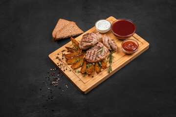 Fototapeta na wymiar Grilled meat steak with fried potato, greens, garlic, sauces and bread served on wooden cutting board. Black wooden textured background, copy space