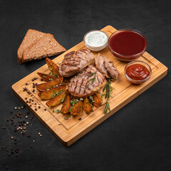Grilled meat steak with fried potato, greens, garlic, sauces and bread served on wooden cutting board. Black wooden textured background, copy space, square format.