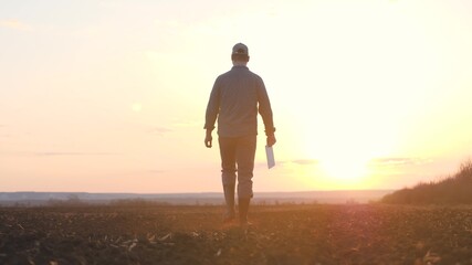 a farmer walks with a tablet in his hand on the ground at sunset in the sky, a working man agronomist is engaged in land cultivation in the field, it is time to fertilize the land for seedlings of the