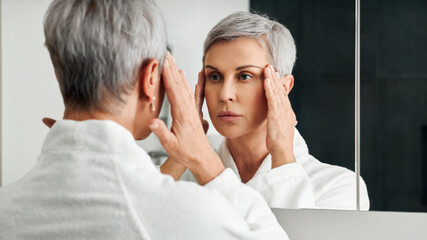 Fototapeta premium Mature woman in a bathroom in front of a mirror touching head with hands