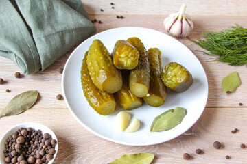 Plate of pickled homemade cucumbers on wooden rustic background. Marinated pickled cucumbers on the white plate with garlic, black pepper and bay leaf. Food concept. Homemade pickled vegetables.