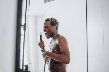 Happy African-American man with a bare torso sings in a glass shower into an imaginary microphone...