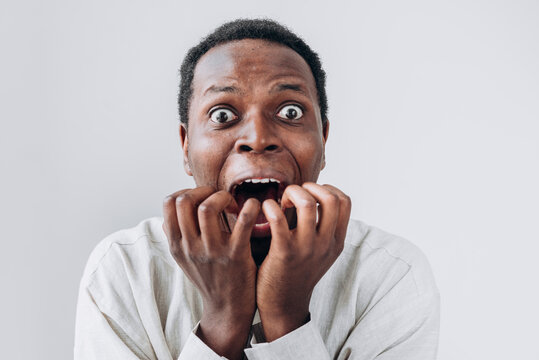 Portrait of a terrified African man staring in horror at the camera and putting his hands to his mouth in a light linen shirt on a white background. Very strong surprise or fright, horror in the eyes.