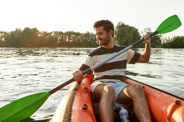 Excited young man smiling while kayaking in a lake, surrounded by peaceful nature scene on a summer afternoon