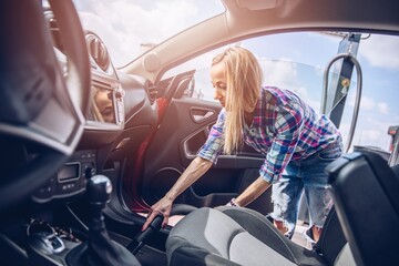 A blond woman wearing a plaid shirt cleans the car inside.