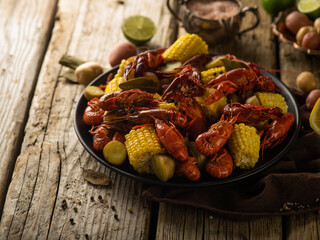 Boiled corn with crayfish. Black plate on a wooden table. There is a gravy bowl with sauce, lime, garlic next to it. Bright paints, dark background. Close-up.