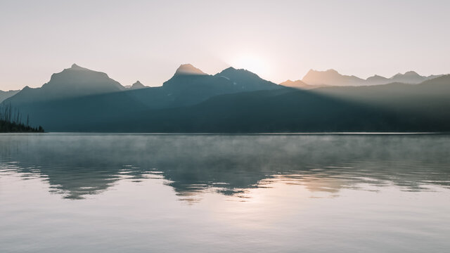 Panoramic image of the sun and sunlight bursting through behind the peaks of the Rocky Mountains with foggy lake reflections on Lake McDonald in Glacier National Park, Montana, USA.