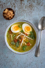 Top down view of a single bowl of Soto Ayam (translation: Indonesian chicken soup) with a spoon on a textured blue background