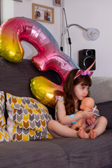 A Cute Baby Girl Sitting On A Sofa With A Colourful Balloon And A Doll