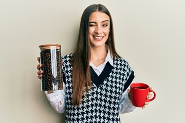 Beautiful brunette young woman holding jar with coffee beans and drinking coffee cup winking...