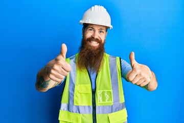 Redhead man with long beard wearing safety helmet and reflective jacket approving doing positive gesture with hand, thumbs up smiling and happy for success. winner gesture.