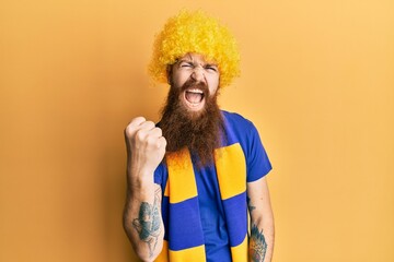 Redhead man with long beard football hooligan cheering game wearing funny wig angry and mad raising...
