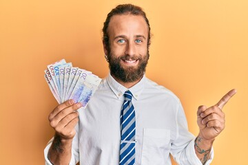 Handsome man with beard and long hair holding swedish krona banknotes smiling happy pointing with hand and finger to the side
