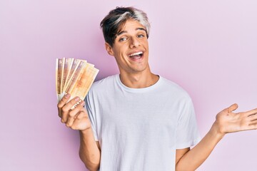 Young hispanic man holding 500 norwegian krone banknotes celebrating achievement with happy smile and winner expression with raised hand