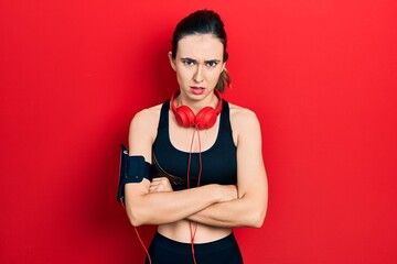 Young hispanic girl wearing gym clothes and using headphones skeptic and nervous, disapproving...