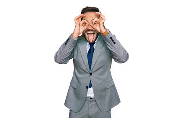 Middle age man wearing business clothes doing ok gesture like binoculars sticking tongue out, eyes looking through fingers. crazy expression.