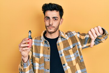 Young hispanic man holding electronic cigarette with angry face, negative sign showing dislike with...