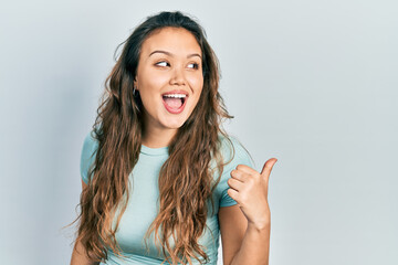 Young hispanic girl wearing casual shirt pointing thumb up to the side smiling happy with open mouth