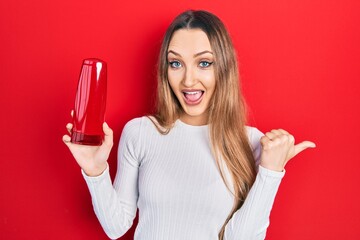 Naklejka premium Young blonde girl holding shampoo bottle pointing thumb up to the side smiling happy with open mouth