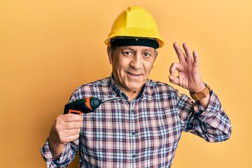 Handsome senior man with grey hair holding screwdriver wearing hardhat doing ok sign with fingers, smiling friendly gesturing excellent symbol