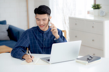 Young asian man talking phone and work from home with laptop computer, freelance male writing on notebook and speak on smartphone, stay home, domestic life, business and communication concept.
