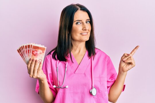 Middle age brunette doctor woman holding 20 shekels banknotes smiling happy pointing with hand and finger to the side