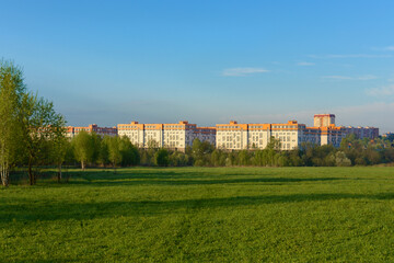 Fototapeta na wymiar Panorama a park scene with a green grass field, a tree plant and a multi-storey residential complex against a blue sky