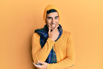 Handsome hispanic man wearing wool hat and winter scarf looking confident at the camera smiling with crossed arms and hand raised on chin. thinking positive.