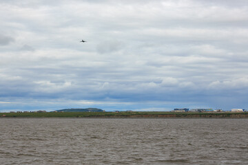 Summer Arctic landscape. A passenger plane is flying in the cloudy sky. Anadyr airport building in the distance. View of the coast of the Anadyr estuary. Travel to the Far North of Russia. Chukotka.