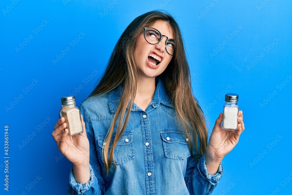 Wall mural Young caucasian woman holding salt shaker angry and mad screaming frustrated and furious, shouting with anger looking up.