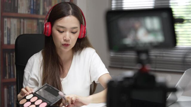 Medium close up shot of Asian young lady talking on cosmetics while holding a makeup palette while recording her video. Woman making a video live tutorial for her cosmetics blog. Cosmetic reviewer.