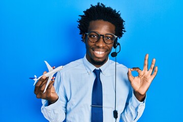 Young african american travel agent man holding plane doing ok sign with fingers, smiling friendly...