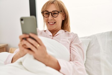 Middle age blonde woman using smartphone lying on the bed at home.