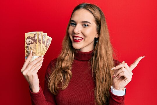 Young blonde woman holding 5000 hungarian forint banknotes smiling happy pointing with hand and finger to the side