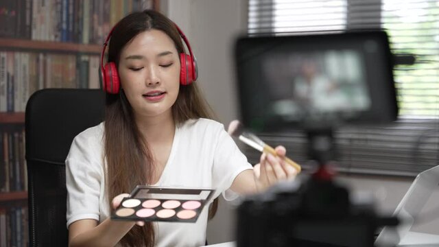Medium close up shot of Asian young lady talking on cosmetics while holding a brush on and demonstrate during record video. Woman making video live tutorial for her cosmetics blog. Cosmetic reviewer.