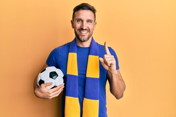 Handsome man with beard football hooligan cheering game holding ball smiling with an idea or question pointing finger with happy face, number one