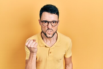 Young hispanic man wearing casual clothes and glasses angry and mad raising fist frustrated and furious while shouting with anger. rage and aggressive concept.