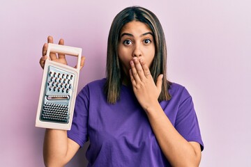 Young latin woman holding grater covering mouth with hand, shocked and afraid for mistake....