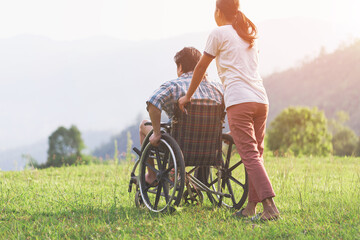 Disabled handicapped man in wheelchair and care helper walking on mountain park. Rear view