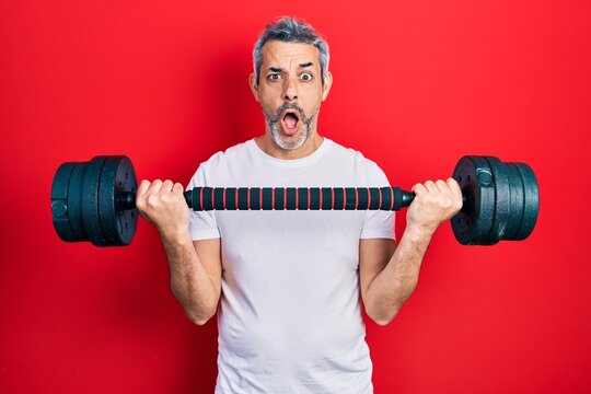 Handsome middle age man with grey hair wearing sportswear using dumbbells afraid and shocked with surprise and amazed expression, fear and excited face.