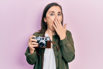 Middle age hispanic woman holding vintage camera covering mouth with hand, shocked and afraid for mistake. surprised expression