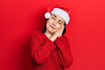 Middle age hispanic woman wearing christmas hat sleeping tired dreaming and posing with hands together while smiling with closed eyes.
