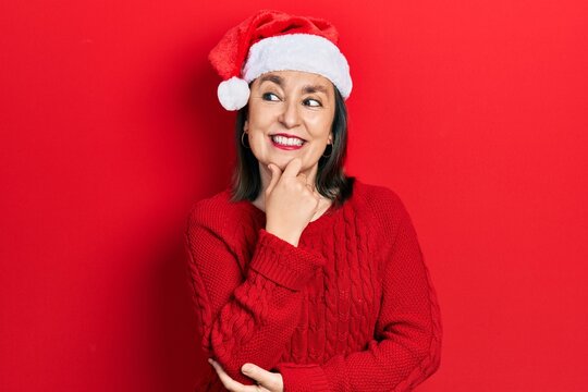 Middle age hispanic woman wearing christmas hat with hand on chin thinking about question, pensive expression. smiling with thoughtful face. doubt concept.