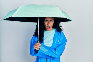 Young hispanic woman with curly hair wearing a raincoat and umbrella in shock face, looking skeptical and sarcastic, surprised with open mouth